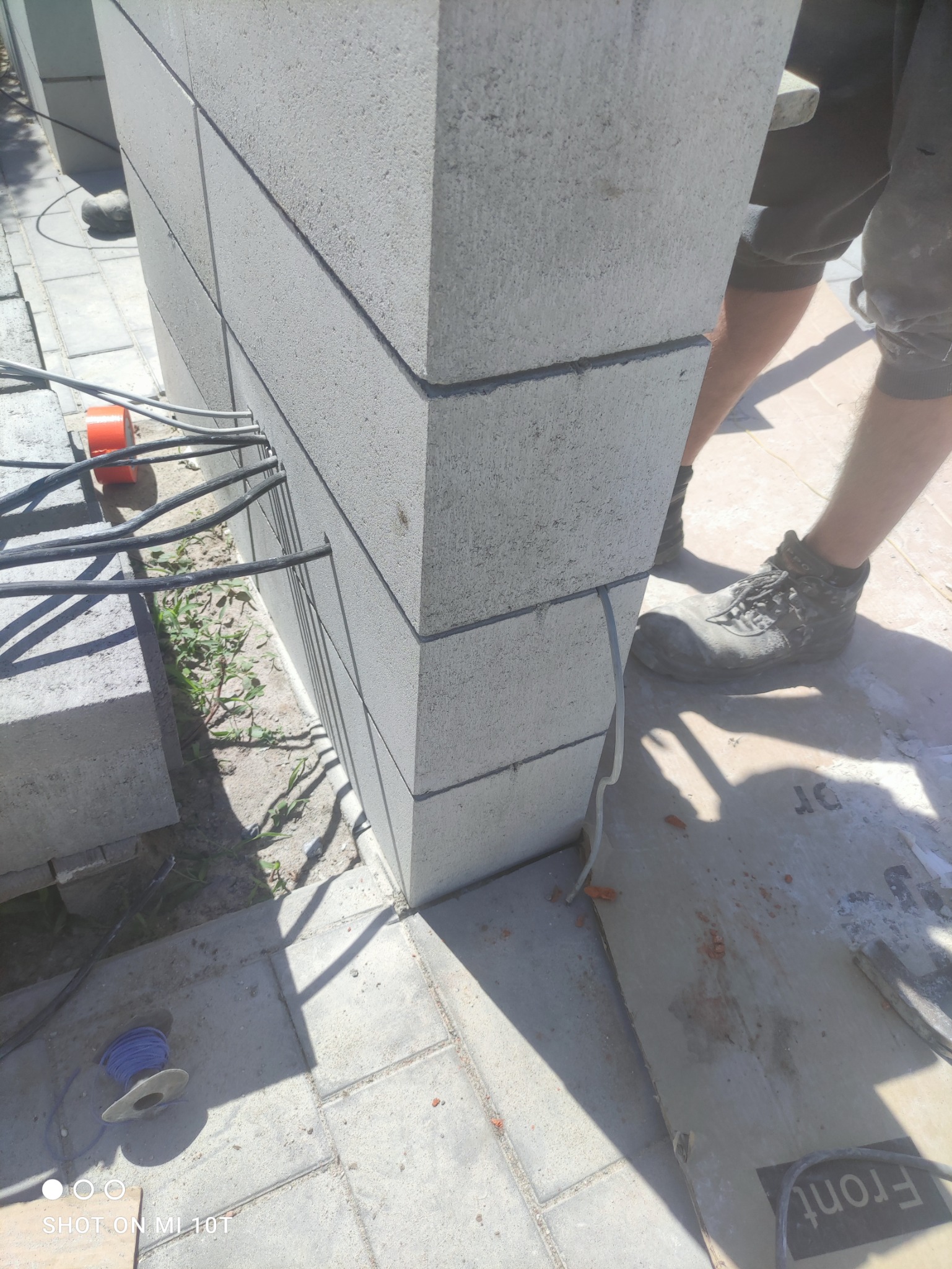 Wznoszenie słupka z szarych bloczków betonowych, widoczne kable elektryczne wystające ze ściany, robocze buty i fragment podłoża z kostki brukowej.
