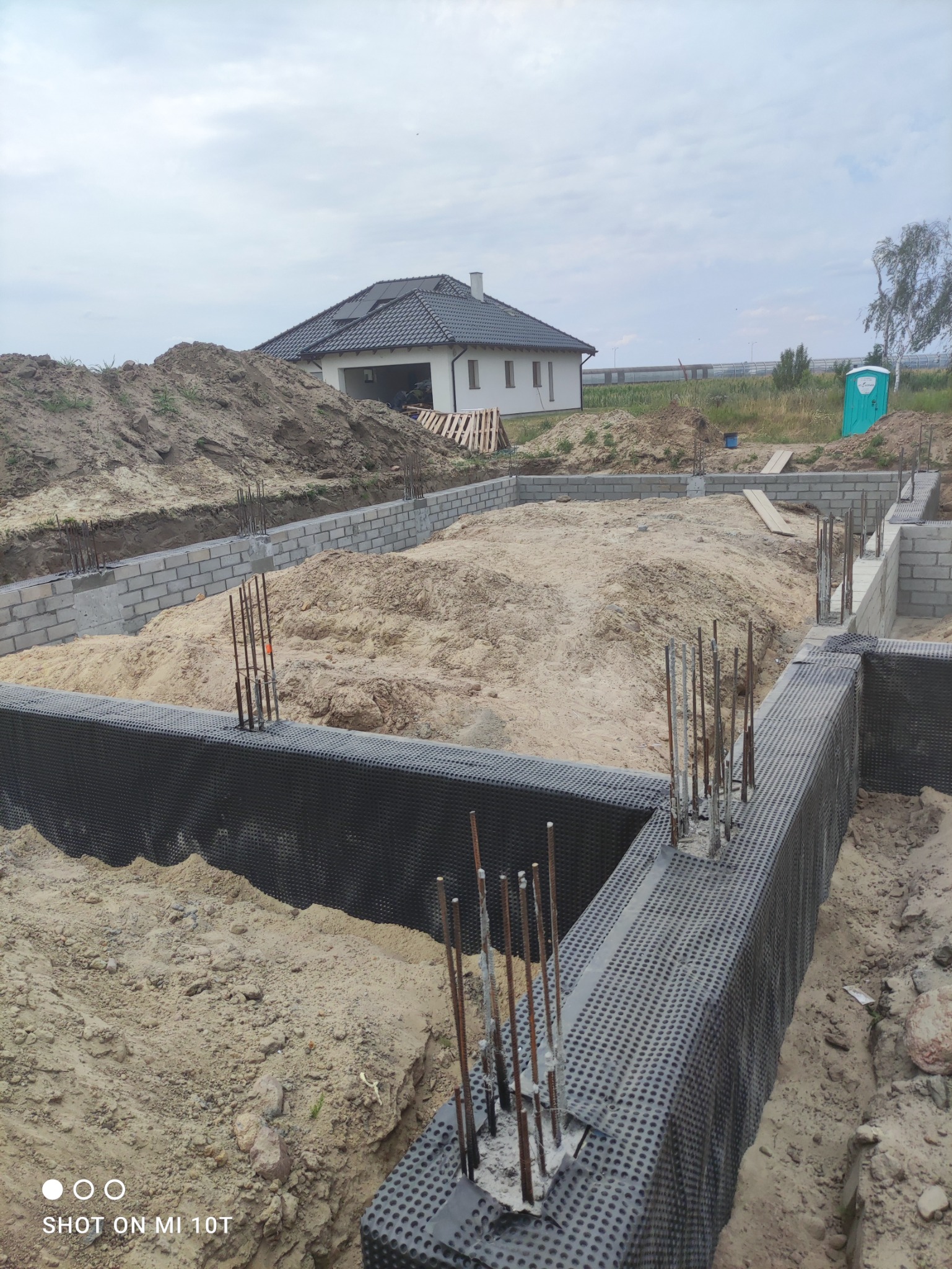 Fundamenty domu jednorodzinnego w trakcie budowy, widoczne zbrojenia i izolacja przeciwwilgociowa, w tle gotowy budynek i przenośna toaleta.