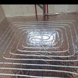 Białe rurki ogrzewania podłogowego ułożone na folii z czerwoną siatką, przymocowane niebieskimi opaskami zaciskowymi, widok z góry na surową podłogę w trakcie montażu.