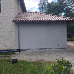 Budynek z nową, gładką, szarą elewacją, kontrastującą z kamienną ścianą i brązowymi rynnami oraz dachówką ceramiczną, na tle zieleni.