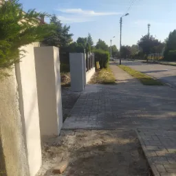 Fragment ogrodzenia z betonowych słupków i metalowych przęseł, świeżo pomalowany na biało i szaro, widok na ulicę i chodnik w słoneczny dzień.