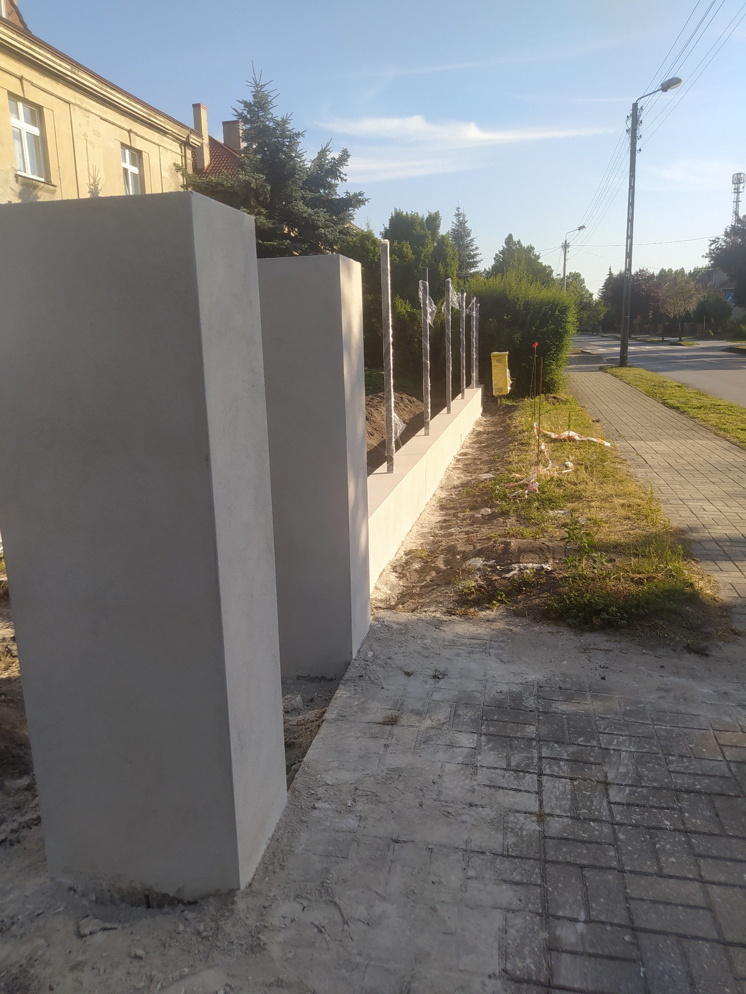 Nowo wybudowany, niski murek z betonowych słupków i metalowych przęseł, zabezpieczonych folią, oddzielający posesję od chodnika i ulicy.