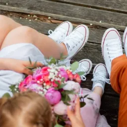 Rodzina siedzi na drewnianym pomoście w białych trampkach Converse, mała dziewczynka trzyma bukiet różowych kwiatów.