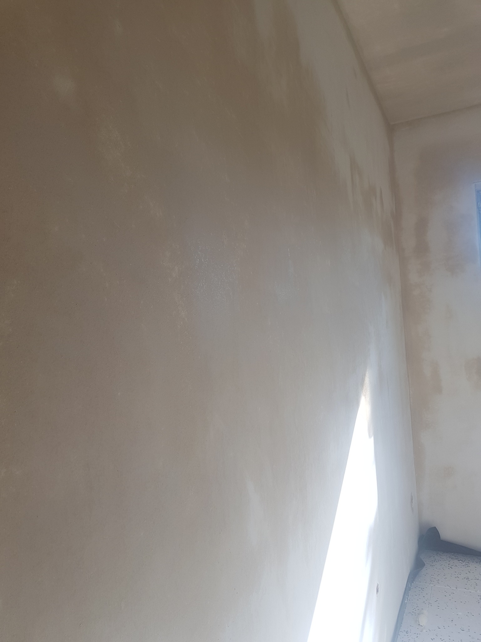 Świeżo nałożona, wilgotna gładź na ścianie w rogu pomieszczenia, z widocznymi smugami i nierównościami przed wyschnięciem, oświetlona ostrym światłem słonecznym wpadającym przez okno.