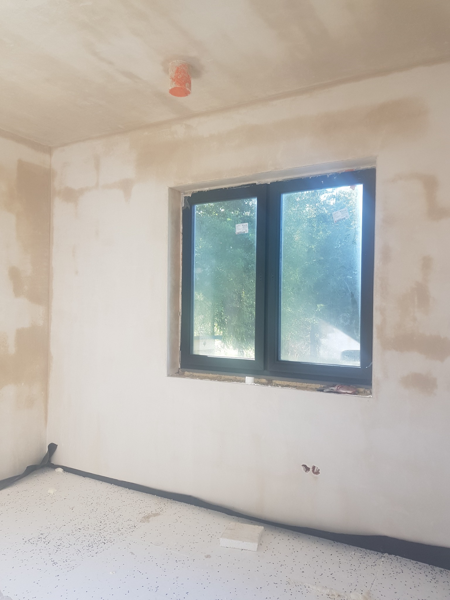 Surowe wnętrze pokoju w trakcie prac wykończeniowych: ściany z częściowo nałożoną gładzią, okno z widokiem na zieleń, zabezpieczona podłoga, widoczne puszki elektryczne pod sufitem.