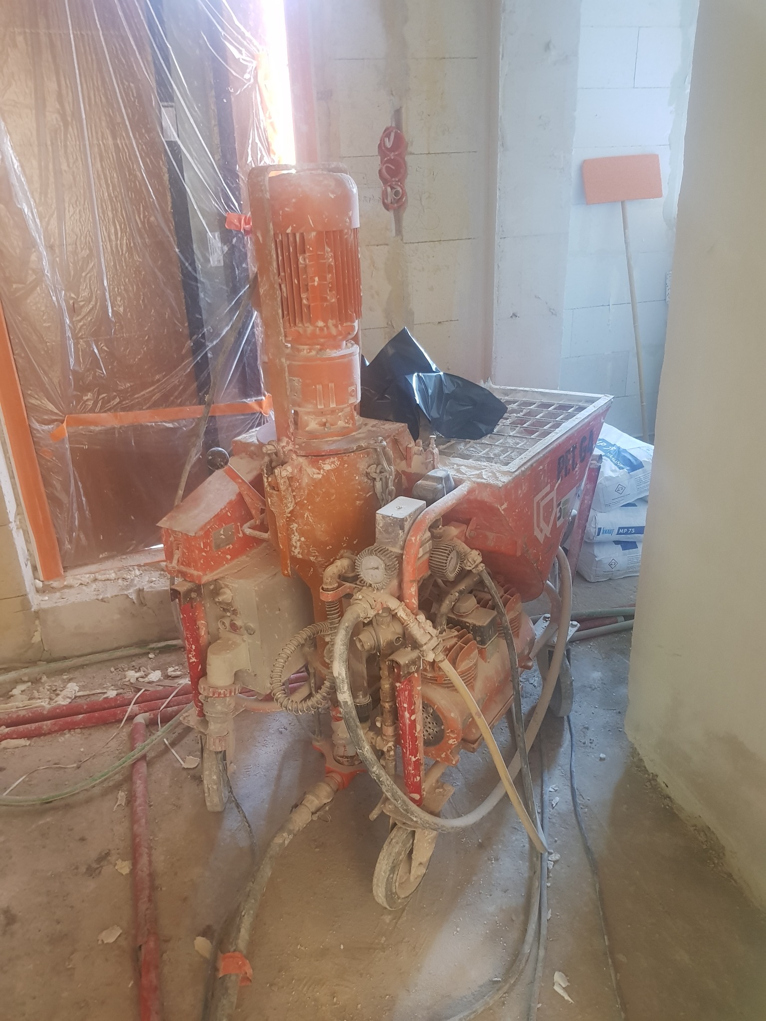 Pomarańczowa maszyna do nakładania gładzi lub tynku, pokryta resztkami materiału, na tle zabezpieczonego folią okna i worków z gładzią.