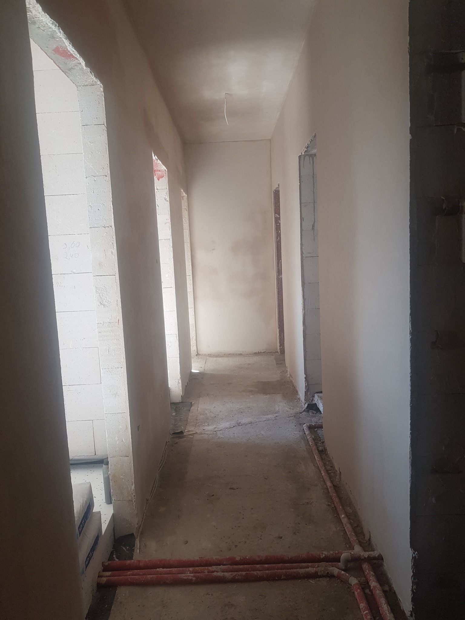 Korytarz w surowym stanie po nałożeniu gładzi, widoczne worki z zaprawą na podłodze, wystające rury i niedokończone ościeżnice.