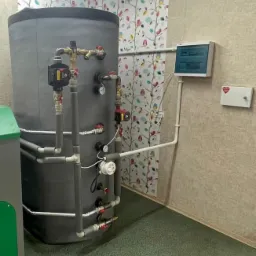 Szary zbiornik akumulacyjny z izolacją i licznymi przyłączami rur, manometrami i pompami obiegowymi w pomieszczeniu z tapetą w kwiatowe wzory i skrzynką elektryczną na ścianie.