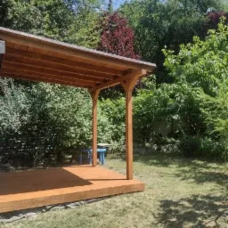 Drewniana konstrukcja altany z brązowym dachem i podłogą, widoczna z boku na tle zielonego ogrodu z drzewami i krzewami, mały niebieski stolik w tle.
