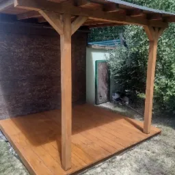 Prosta drewniana konstrukcja z zadaszeniem i drewnianym podestem, świeżo po impregnacji, widok z poziomu gruntu, w tle budynek gospodarczy i zieleń.