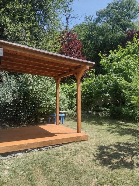 Drewniana konstrukcja altany z brązowym dachem i podłogą, widoczna z boku na tle zielonego ogrodu z drzewami i krzewami, mały niebieski stolik w tle.