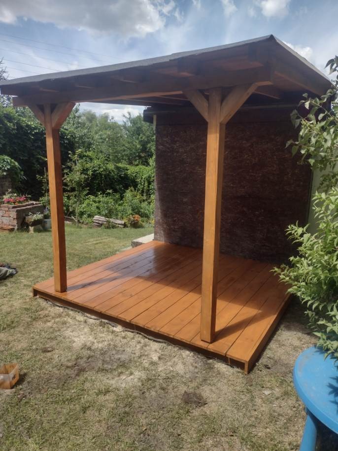 Świeżo polakierowana, drewniana konstrukcja z zadaszeniem w ogrodzie, widoczne nierówności trawnika i ślady po pracy.