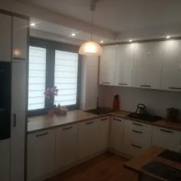 Jasna kuchnia z białymi, błyszczącymi frontami szafek, drewnianym blatem, czarnym zlewem i minimalistycznymi uchwytami, oświetlona wiszącą lampą i punktowymi światłami.