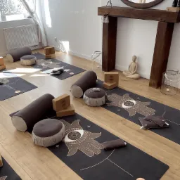 Jasne studio z rozłożonymi matami, wałkami i poduszkami do ćwiczeń, drewnianą atrapą kominka z owalnym lustrem i figurką Buddy.