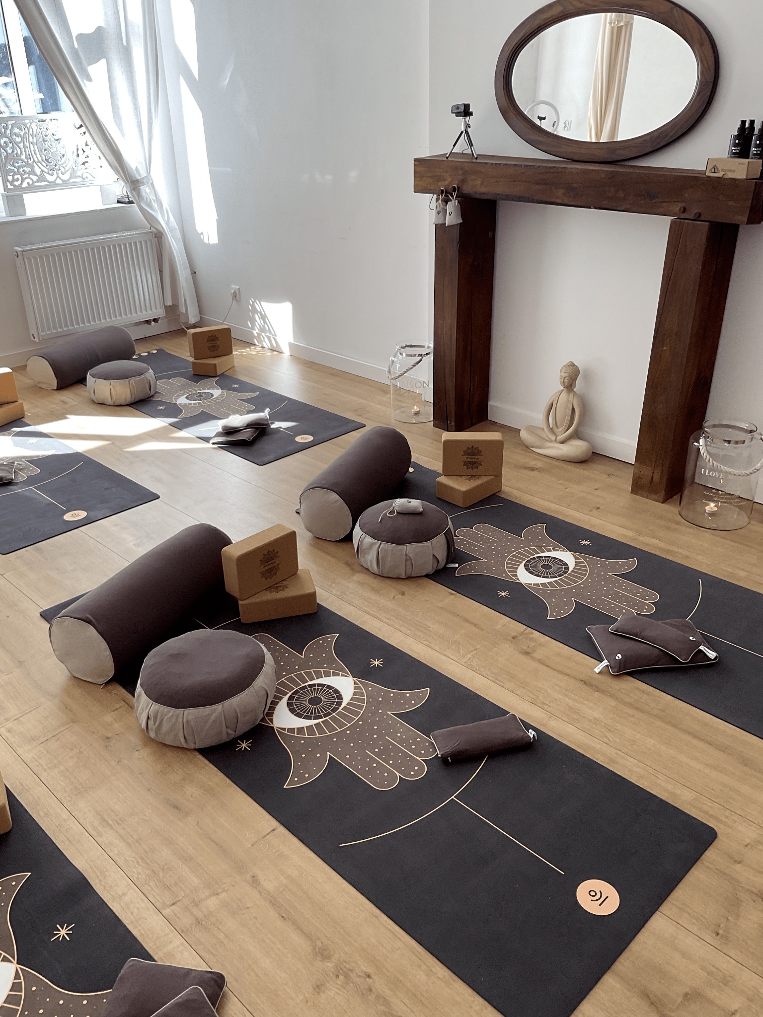 Jasne studio z rozłożonymi matami, wałkami i poduszkami do ćwiczeń, drewnianą atrapą kominka z owalnym lustrem i figurką Buddy.