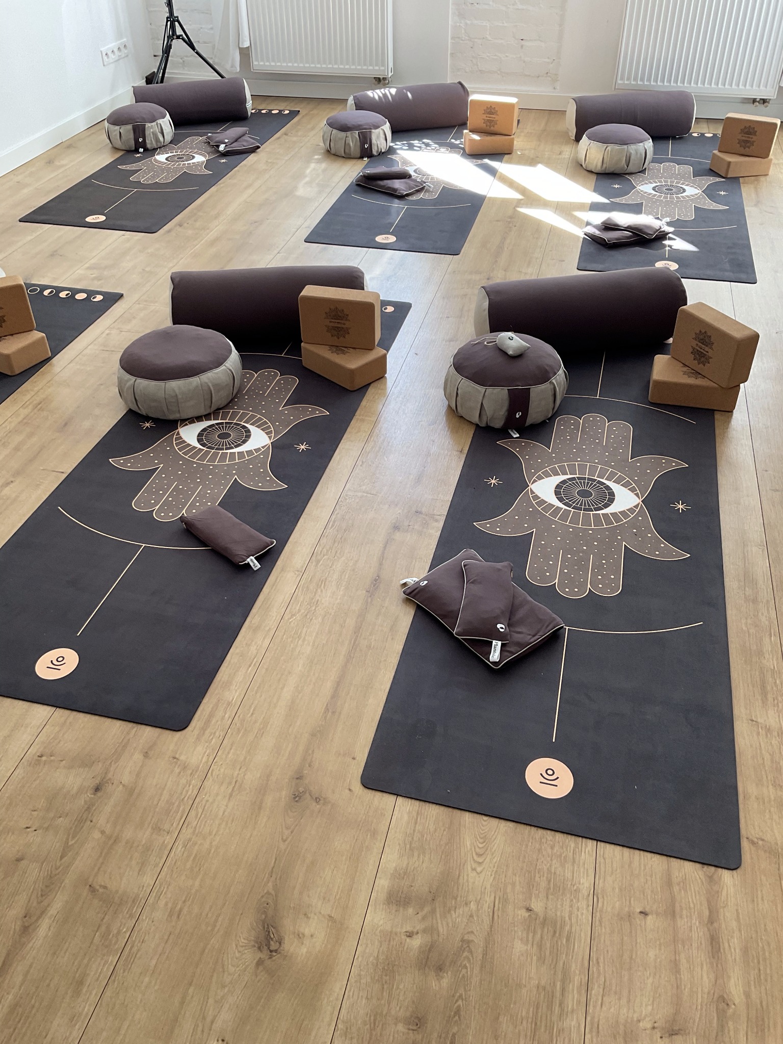Przygotowane stanowiska do jogi: czarne maty z motywem Hamsa, brązowe poduszki do medytacji, wałki i drewniane klocki na jasnej drewnianej podłodze w jasnym pomieszczeniu.