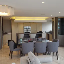 Nowoczesna jadalnia z widokiem na miasto, oświetlona dekoracyjną lampą sufitową i podświetleniem nad blatem kuchennym, widoczne gniazdko elektryczne przy oknie.