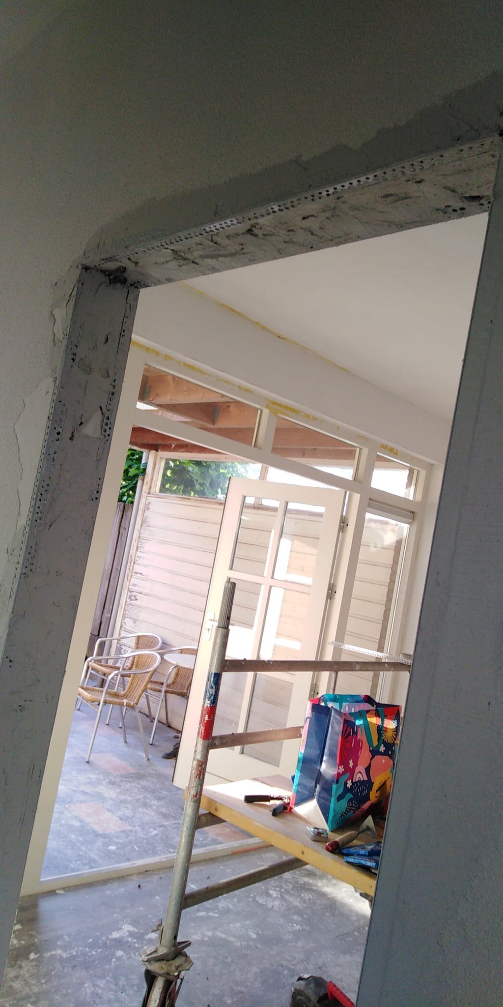 Przygotowania do prac wykończeniowych: widok przez surową futrynę na taras z meblami ogrodowymi, częściowo przeszklone drzwi opierają się o rusztowanie, obok torba na zakupy i narzędzia.