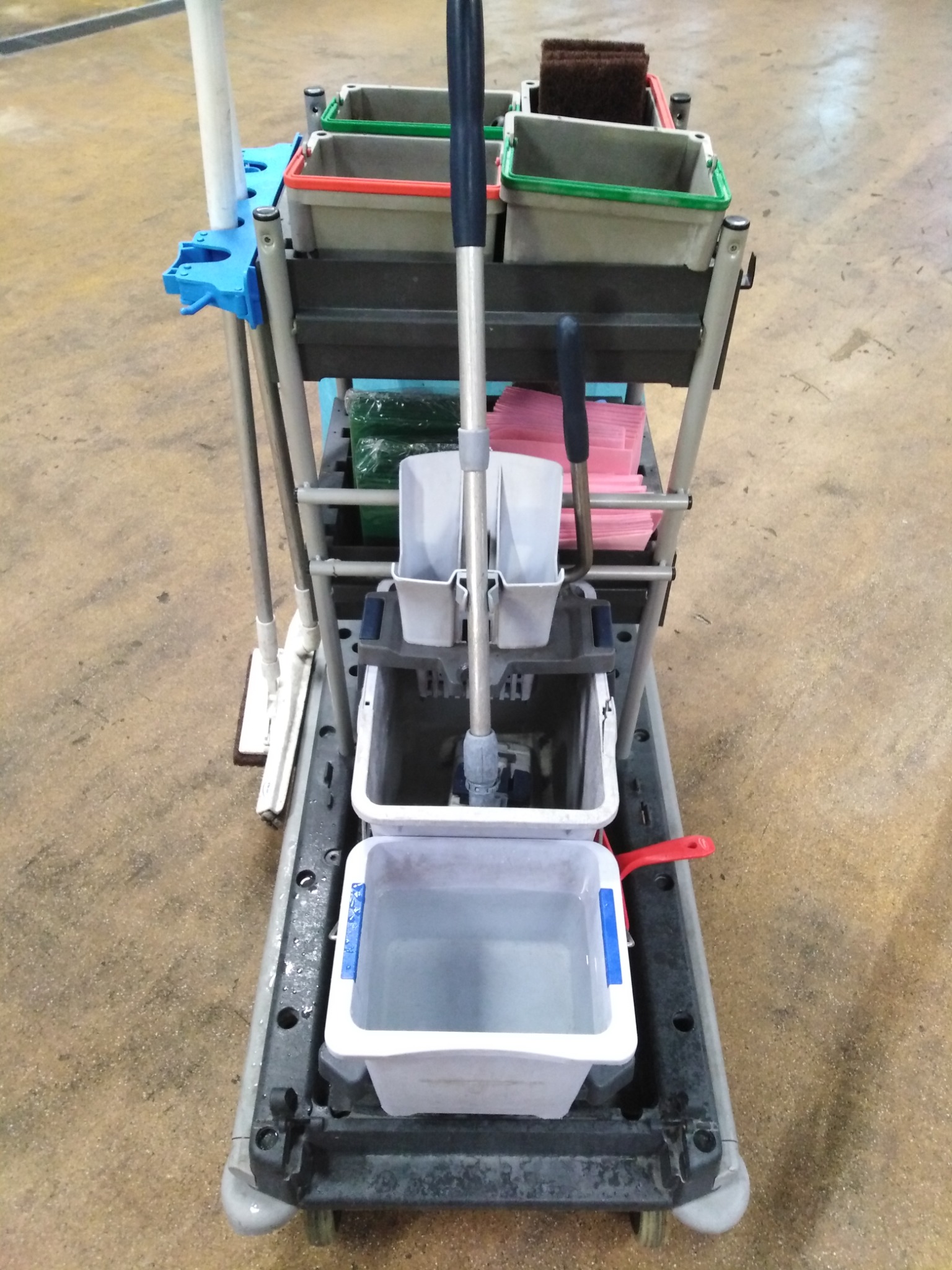 Wielopoziomowy wózek do sprzątania z mopem, wiadrem z wodą, szczotkami i stertą różowych ścierek na tle podłogi z efektem lastryko.