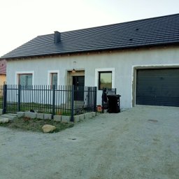 Firma Handlowo Uslugova DEMJANCZYK - Szary dom jednorodzinny z ciemną dachówką i garażem, widoczny z zewnątrz, ogrodzony czarnym metalowym płotem, z fragmentami elewacji wymagającymi wykończenia.