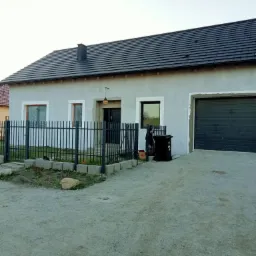 Szary dom jednorodzinny z ciemną dachówką i garażem, widoczny z zewnątrz, ogrodzony czarnym metalowym płotem, z fragmentami elewacji wymagającymi wykończenia.