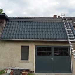 Firma Handlowo Uslugova DEMJANCZYK - Nowy, grafitowy dach z blachodachówki trapezowej na budynku gospodarczym w Kościanie, z widoczną drabiną opartą o dach i stertą starych dachówek przy ścianie.