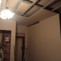 Firma Handlowo Uslugova DEMJANCZYK - Montaż płyt gipsowo-kartonowych na ścianach i suficie, widoczne profile stalowe i wisząca lampa w surowym pomieszczeniu z instalacją grzewczą i drzwiami.