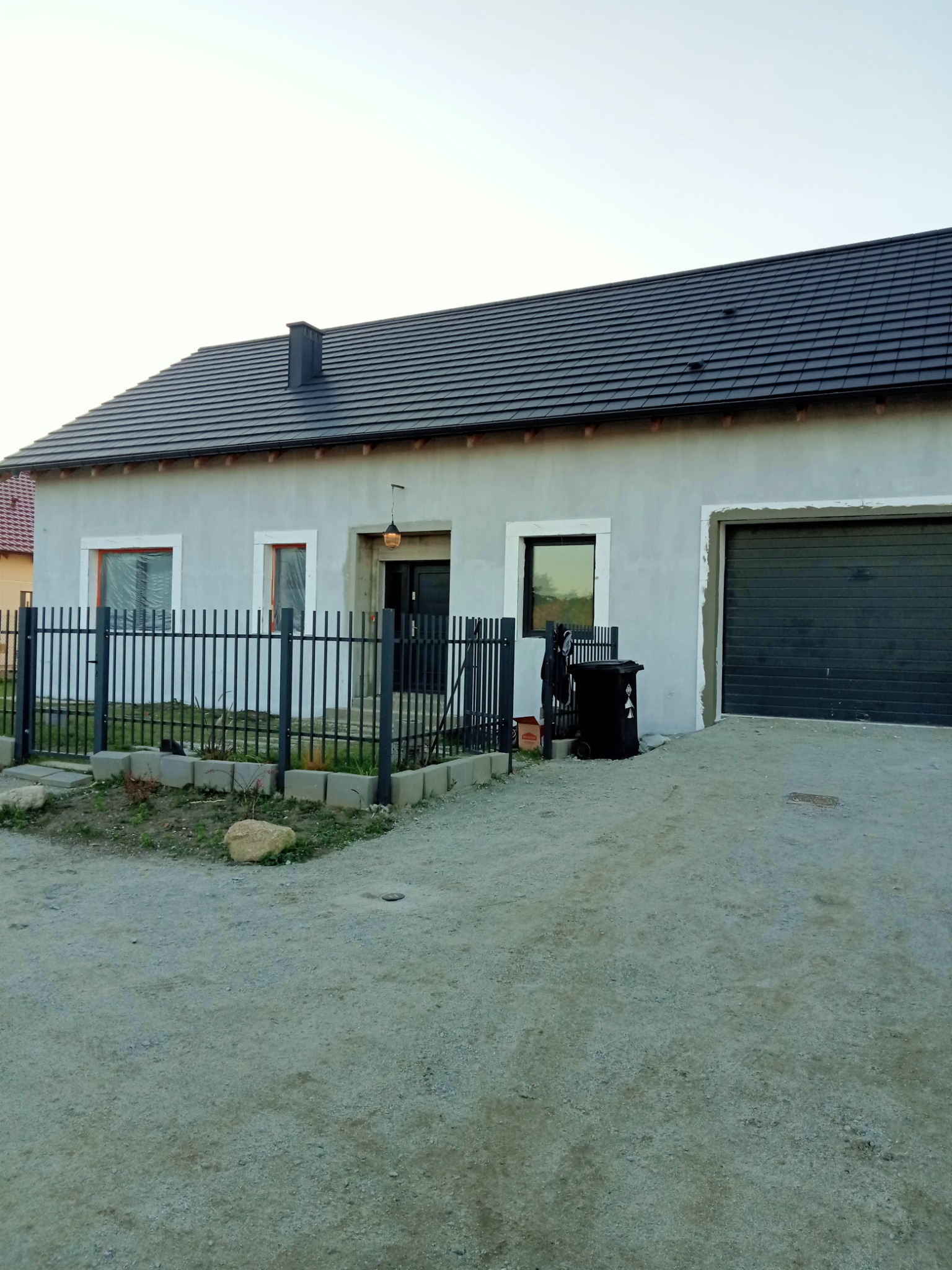 Szary dom jednorodzinny z ciemną dachówką i garażem, widoczny z zewnątrz, ogrodzony czarnym metalowym płotem, z fragmentami elewacji wymagającymi wykończenia.