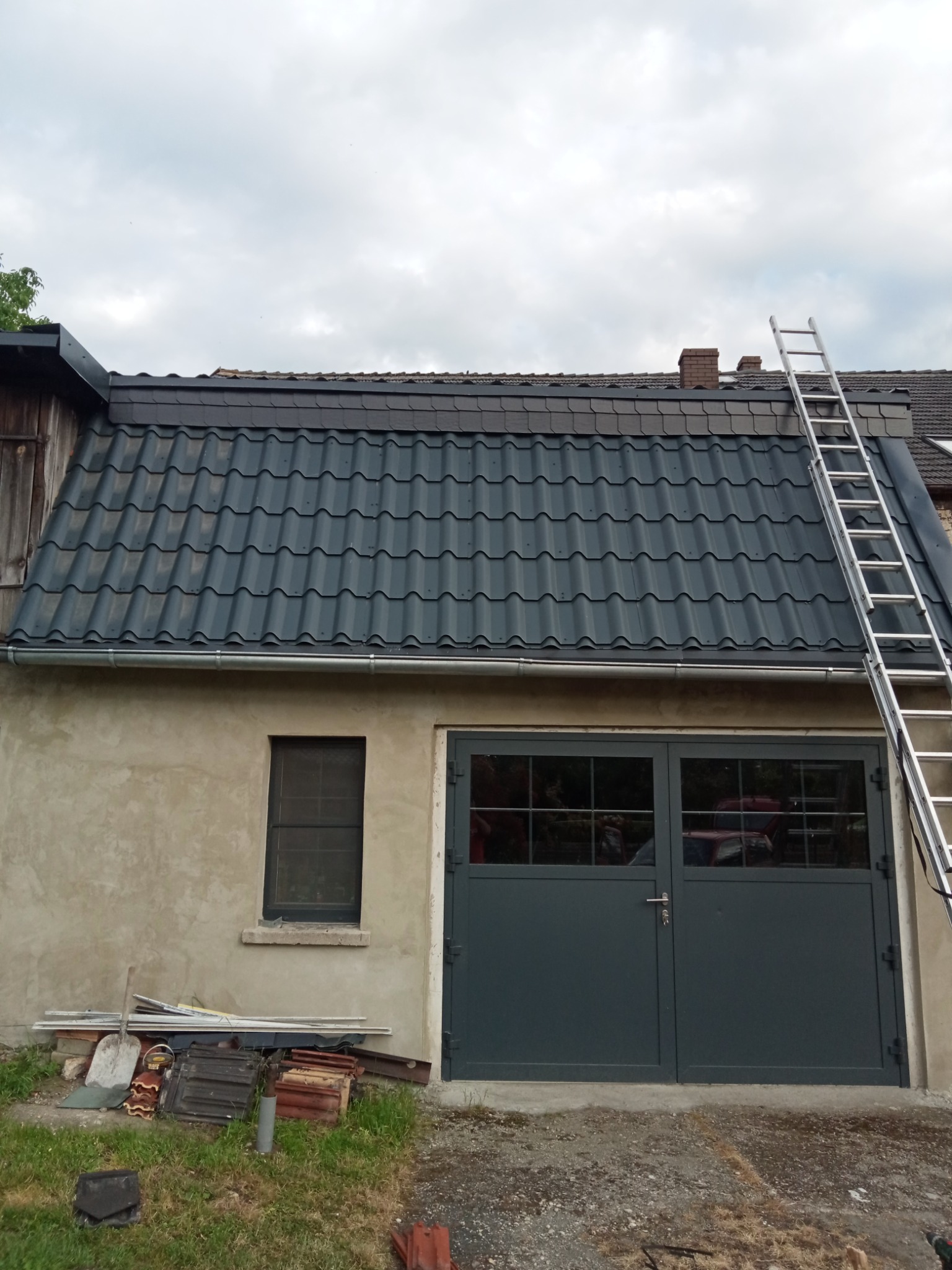 Nowy, grafitowy dach z blachodachówki trapezowej na budynku gospodarczym w Kościanie, z widoczną drabiną opartą o dach i stertą starych dachówek przy ścianie.