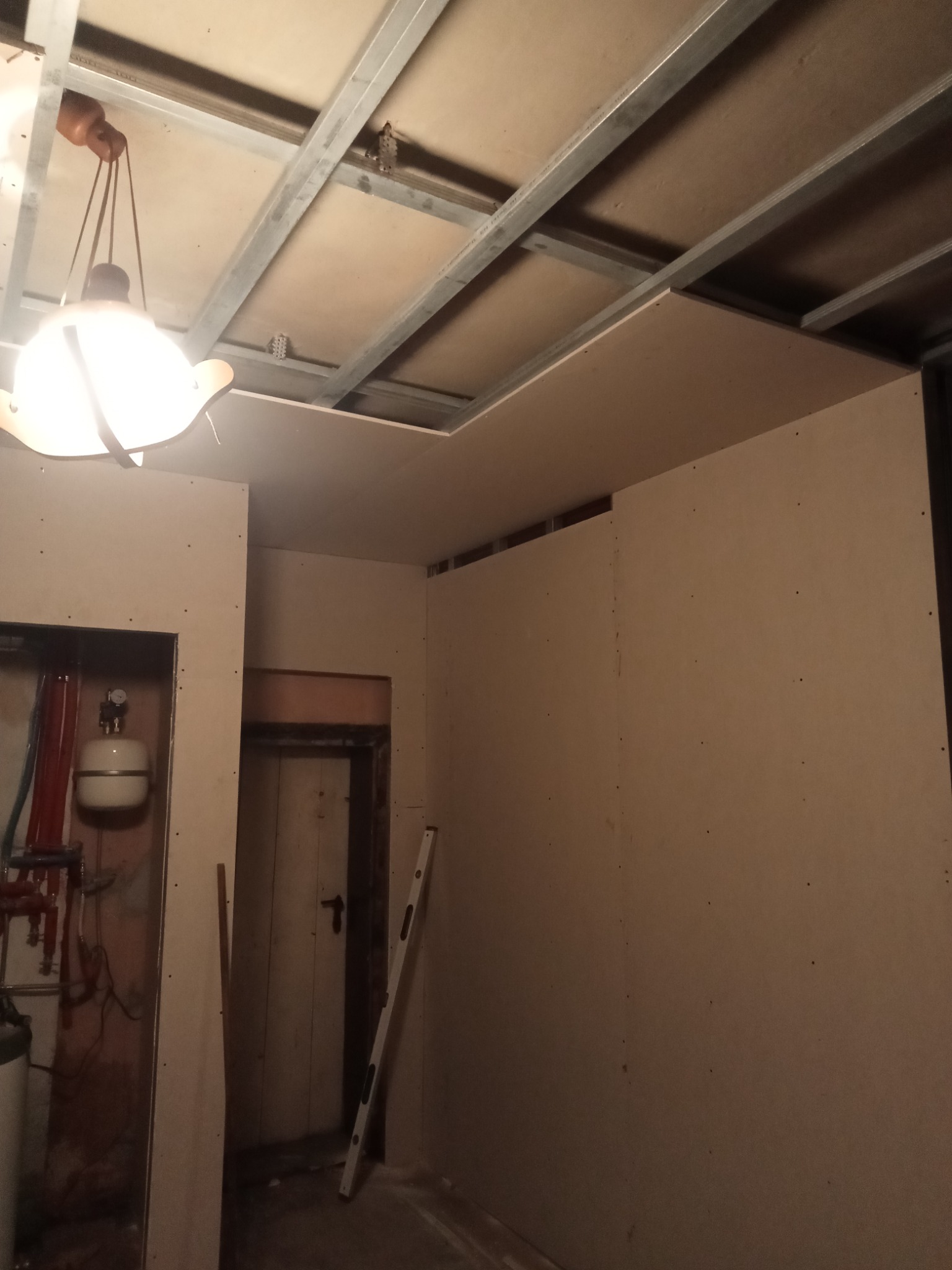 Montaż płyt gipsowo-kartonowych na ścianach i suficie, widoczne profile stalowe i wisząca lampa w surowym pomieszczeniu z instalacją grzewczą i drzwiami.