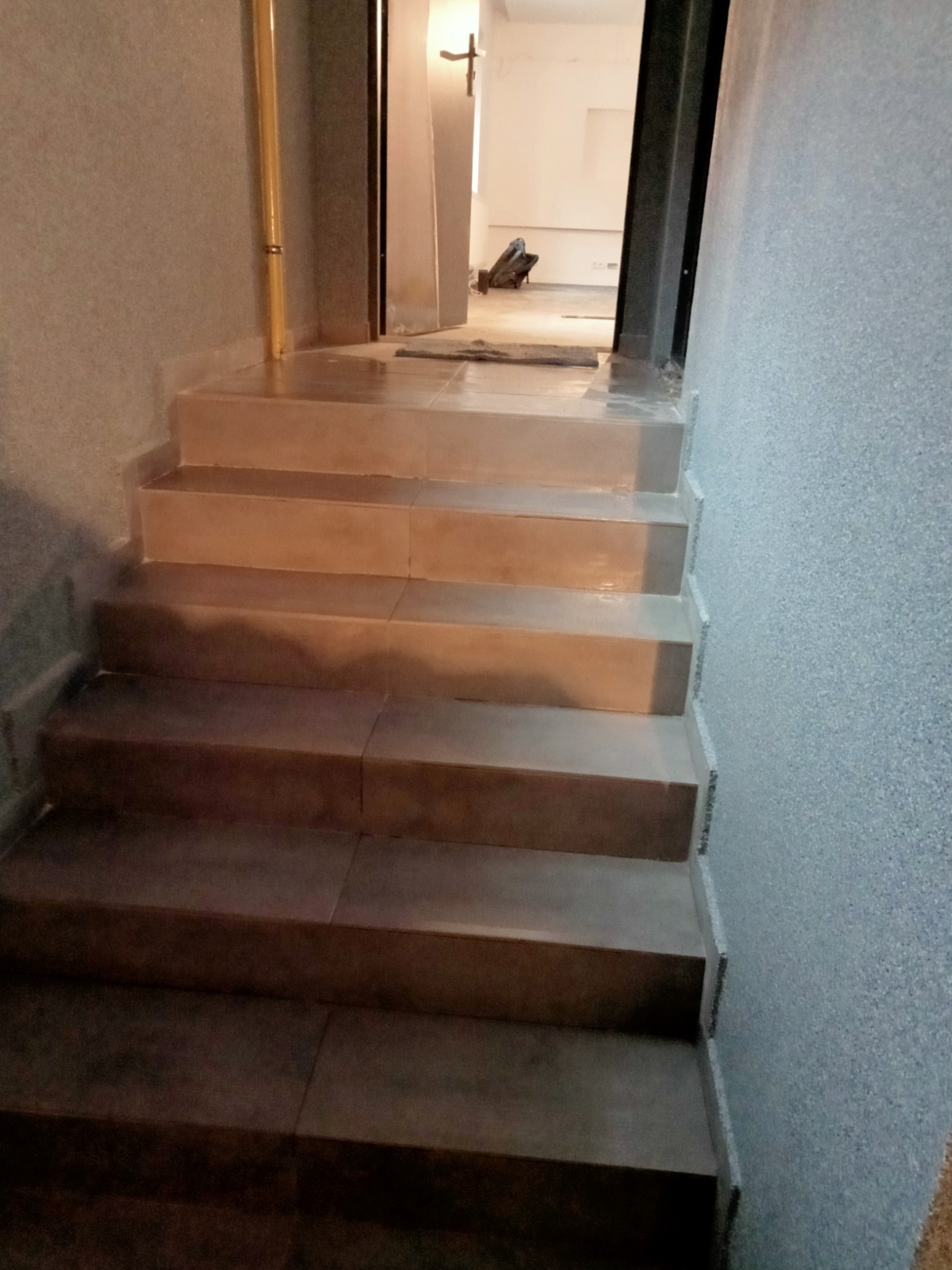 Wykończone schody wewnętrzne z płytek ceramicznych prowadzące do jasnego pomieszczenia, widoczna faktura ściany i fragment rury gazowej w narożniku.