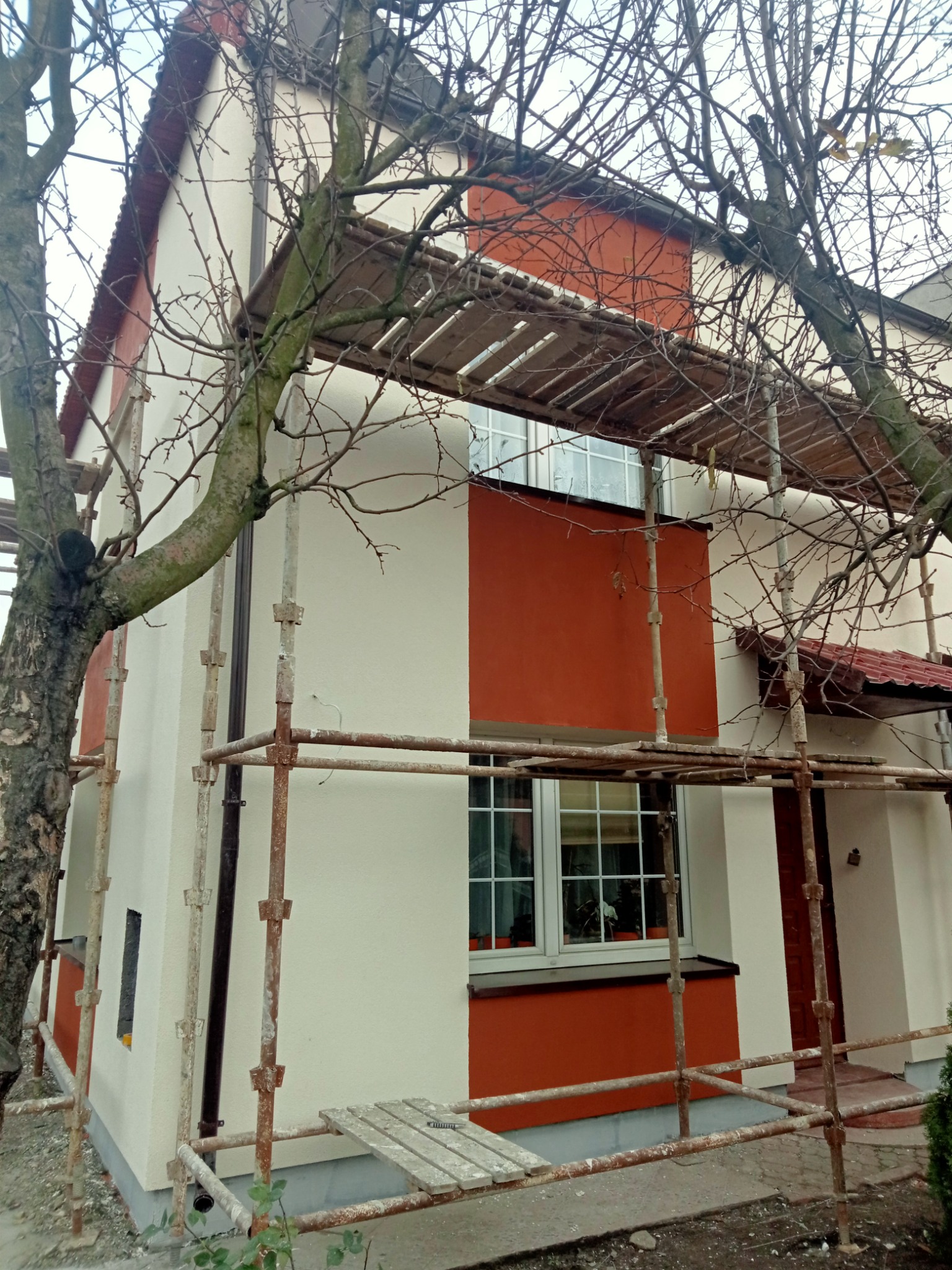 Dwukondygnacyjny dom w trakcie malowania elewacji na biało i pomarańczowo, otoczony metalowymi rusztowaniami i drzewem bez liści.