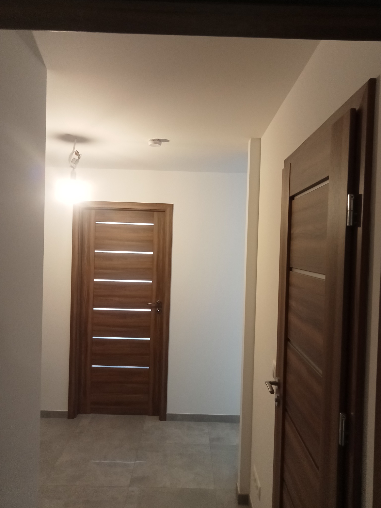 Nowoczesne drzwi wewnętrzne z poziomymi przeszkleniami w minimalistycznym korytarzu z szarą podłogą i białymi ścianami, widoczne niedokończone podłączenie oświetlenia sufitowego.