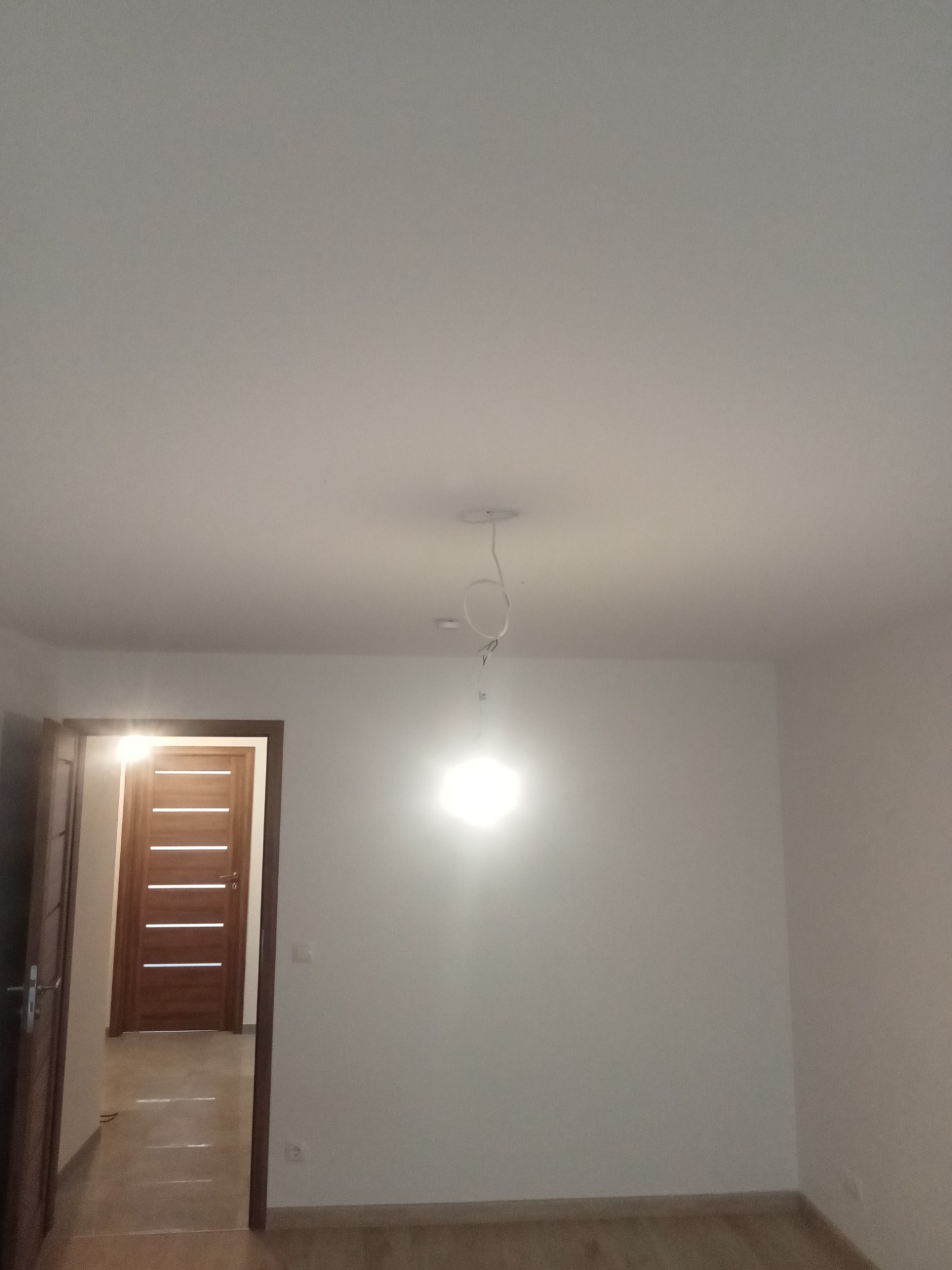 Puste, białe pomieszczenie z drewnianą podłogą i drzwiami wewnętrznymi z poziomymi panelami, widoczne kable wystające z sufitu w miejscu planowanego oświetlenia.