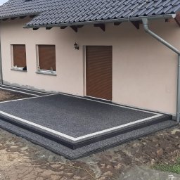 Wiktor Będkowski - Nowoczesny taras z ciemnego kruszywa z białą obwódką, przylegający do beżowej elewacji domu, z widocznym niezakończonym otoczeniem i sprzętem.