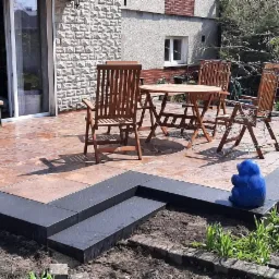 Ukończony taras z kamienną nawierzchnią w Opolu, z ciemnymi obrzeżami i schodami, uzupełniony o drewniane meble ogrodowe i dekoracyjne, niebieskie kule i figurki.