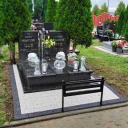 Podw&oacute;jny, czarny nagrobek z granitową płytą, otoczony jasnoszarą kostką brukową i czarną obw&oacute;dką, z metalową ławeczką, świecami i kwiatami, na tle cmentarza z tujami i innymi nagrobkami.