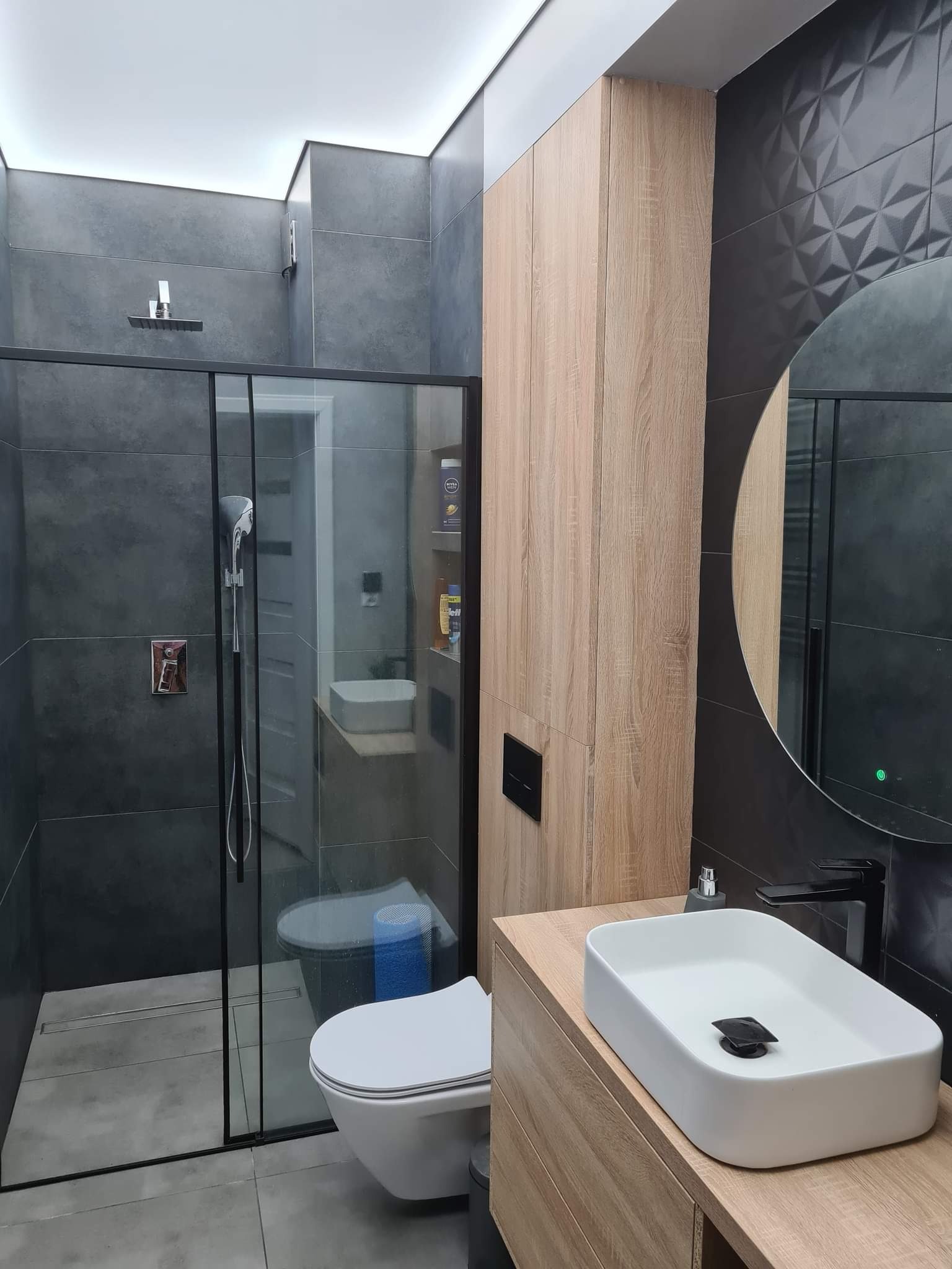 Nowoczesna łazienka z szarymi płytkami, kabiną prysznicową typu walk-in z czarnymi profilami, wiszącą toaletą, umywalką nablatową na drewnianej szafce oraz okrągłym lustrem z podświetleniem LED.