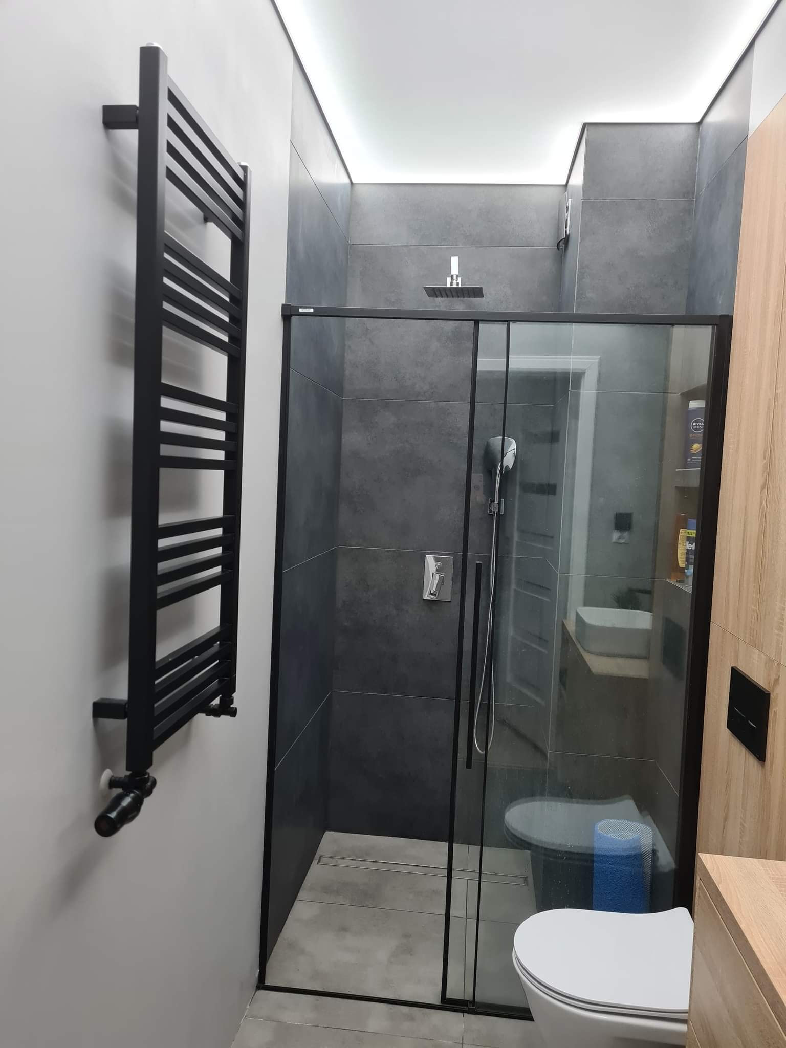 Nowoczesna łazienka z szarymi płytkami, kabiną prysznicową typu walk-in z czarnymi profilami i grzejnikiem drabinkowym w tym samym kolorze, oświetleniem LED podsufitowym i elementami wykończenia...