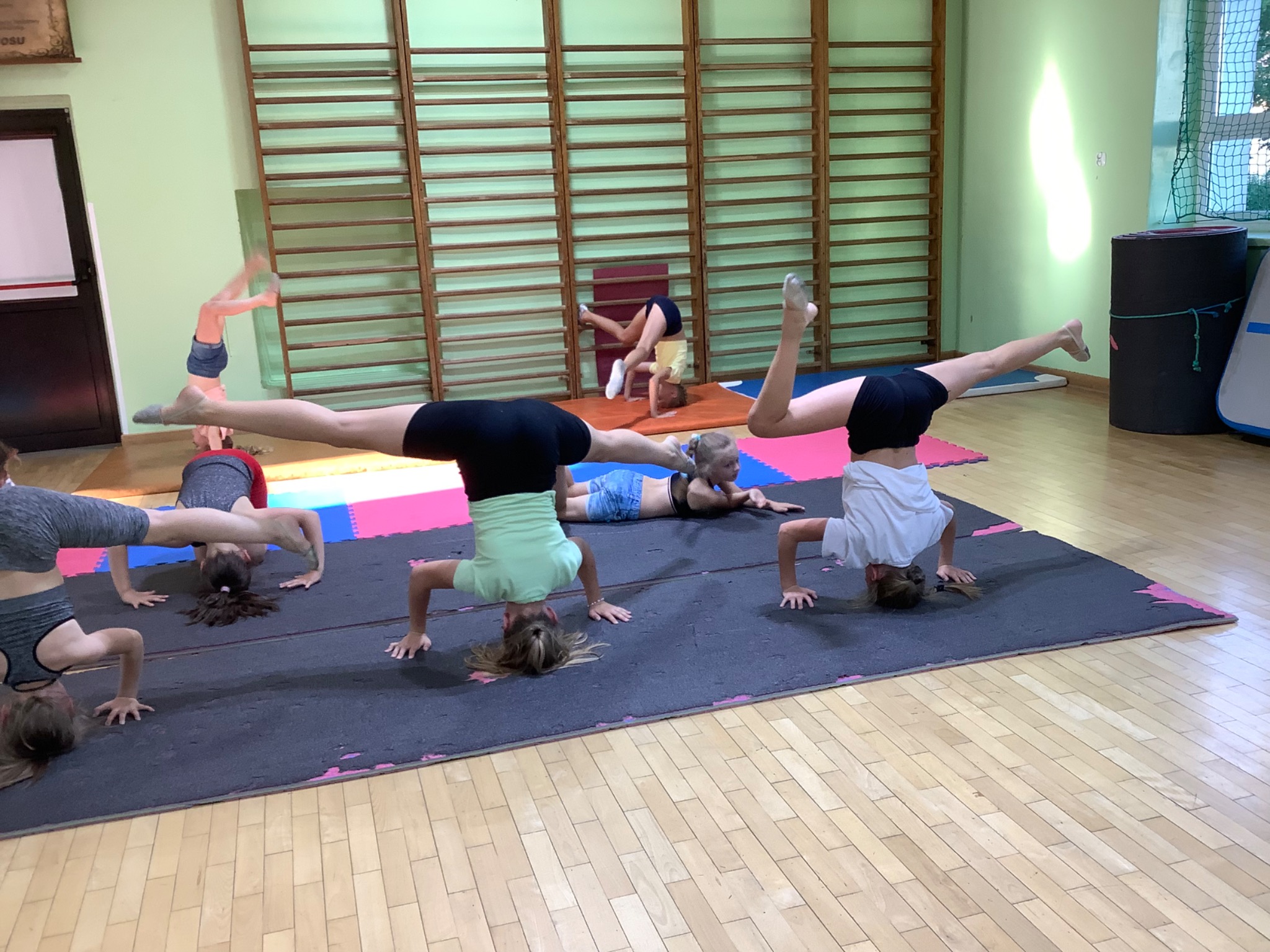 Grupa dzieci ćwiczących stanie na głowie i rozciąganie nóg w sali gimnastycznej z drabinkami na ścianie.