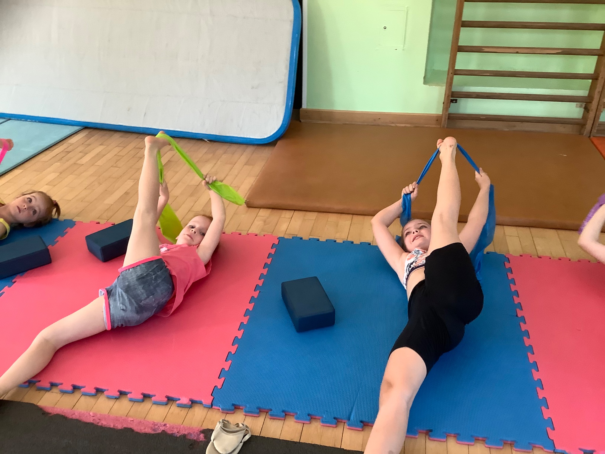 Dziewczynki leżą na kolorowych matach, wykonując ćwiczenia rozciągające z gumami oporowymi, w tle drabinki gimnastyczne i materac.