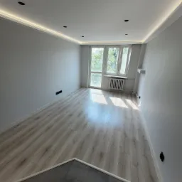 Pomalowane na szaro wnętrze pokoju z panelami podłogowymi w odcieniach drewna i szarą strefą wejściową, oświetlenie sufitowe LED, okno balkonowe z grzejnikiem.