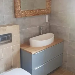 Wykończona łazienka w Krakowie z szarymi płytkami imitującymi drewno na ścianach i podłodze ułożonymi w jodełkę, wisząca toaleta, szafka pod umywalką z drewnianym blatem i owalną umywalką...