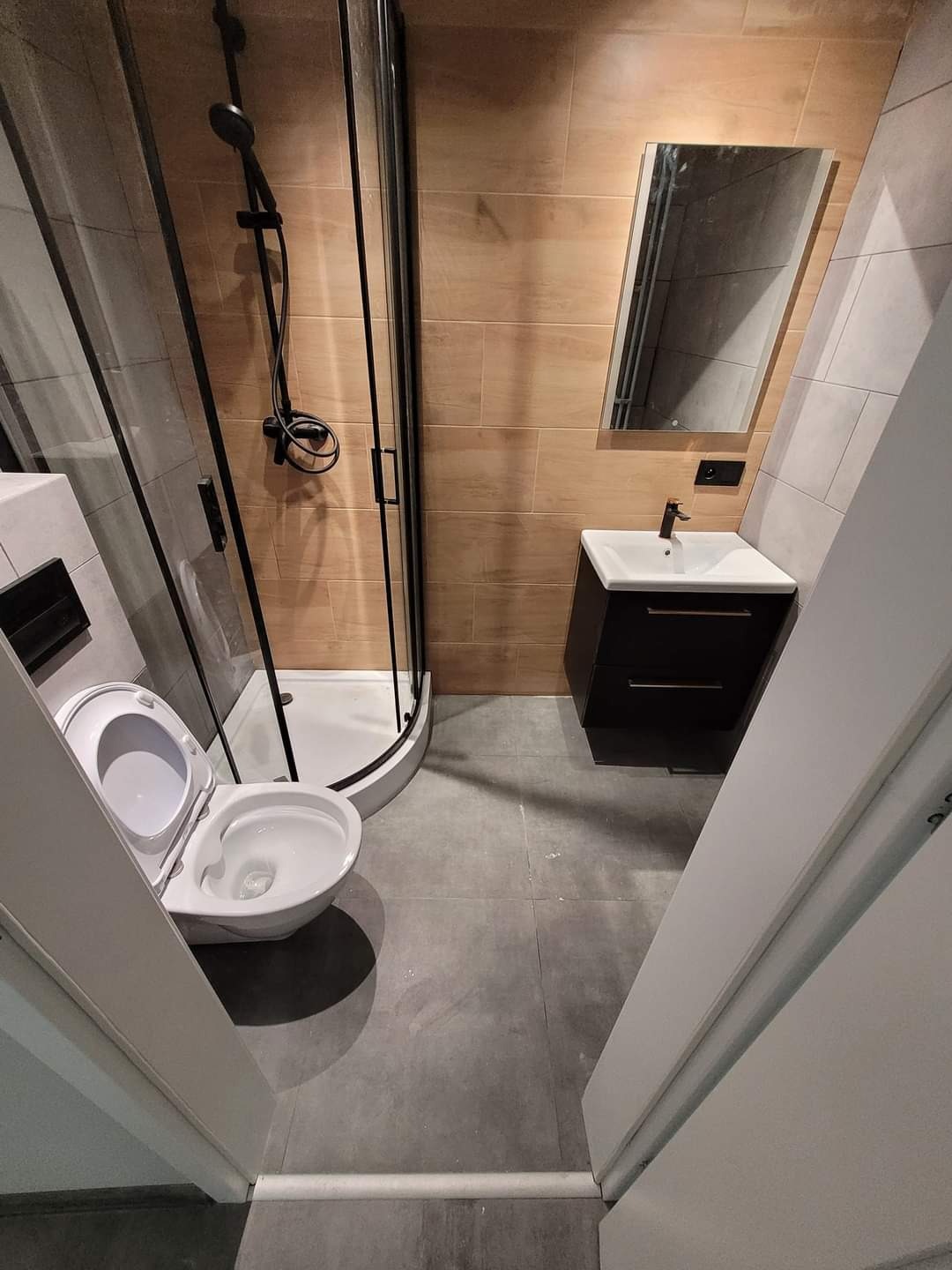 Nowoczesna łazienka po renowacji z kabiną prysznicową z czarnymi ramami, wiszącą toaletą, umywalką z szafką w kolorze ciemnego drewna i podświetlanym lustrem, wykończona płytkami w odcieniach beżu...