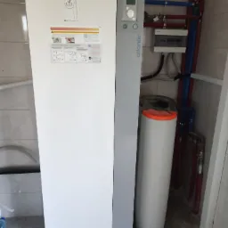 Biała pompa ciepła Atlantic Alfea z wyświetlaczem i zbiornikiem buforowym, widoczne rury instalacji w kolorze niebieskim i czerwonym, na ścianie instrukcja obsługi.