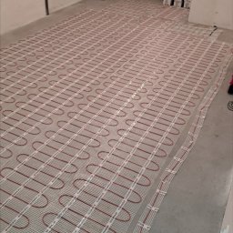 JGinstal usługi elektryczne Jakub Garus - Rozłożona mata grzewcza z czerwonymi przewodami na szarej podłodze w pomieszczeniu, przygotowana do zalania wylewką. W tle worki z zaprawą i kartony.