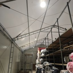 JGinstal usługi elektryczne Jakub Garus - Wnętrze magazynu z metalową konstrukcją dachu, oświetleniem punktowym i regałami wypełnionymi materiałami tekstylnymi w foliowych opakowaniach.