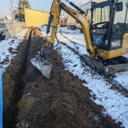 JGinstal usługi elektryczne Jakub Garus - Żółta koparka CAT 301.8 wykopuje rów w ziemi, w którym widać niebieskie rury. Prace ziemne na zewnątrz budynku w zimowej scenerii. Wykop pod przyłącze elektryczne.