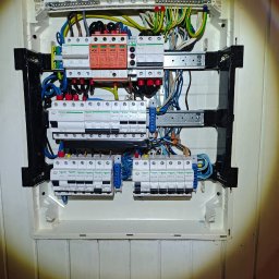 JGinstal usługi elektryczne Jakub Garus - Rozdzielnia elektryczna z zabezpieczeniami przepięciowymi i wyłącznikami różnicowoprądowymi, zamontowana w białej obudowie na ścianie z boazerią.