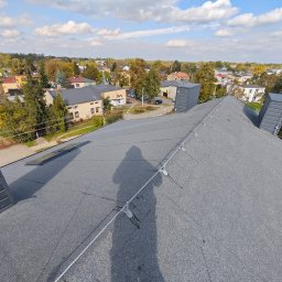 JGinstal usługi elektryczne Jakub Garus - Szary dach z widoczną instalacją odgromową i kominami wentylacyjnymi na tle zabudowy miejskiej w słoneczny dzień. Widoczny cień osoby fotografującej.